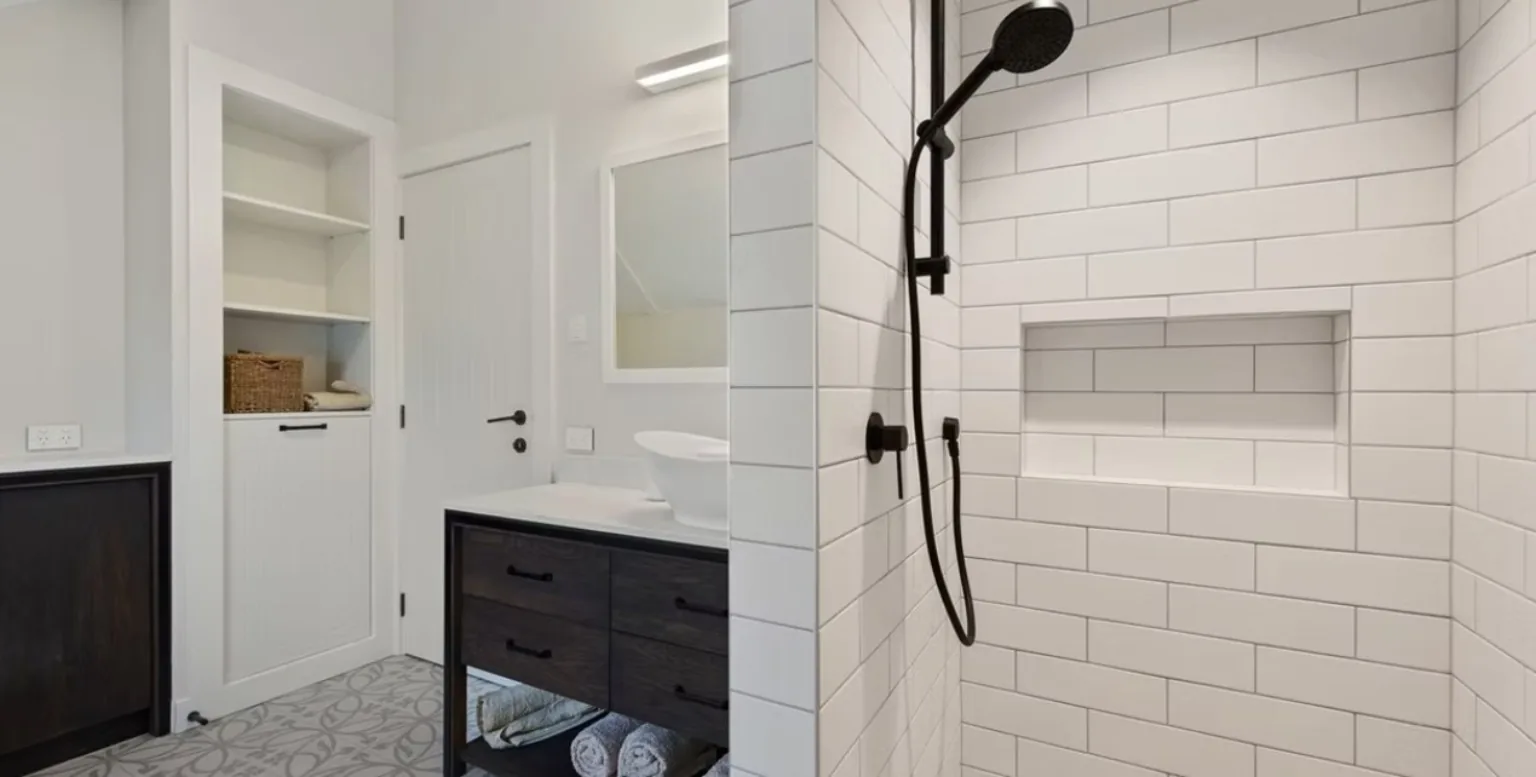A white subway tiled shower with a recessed shelf and black tapware, beside a dark timber vanity